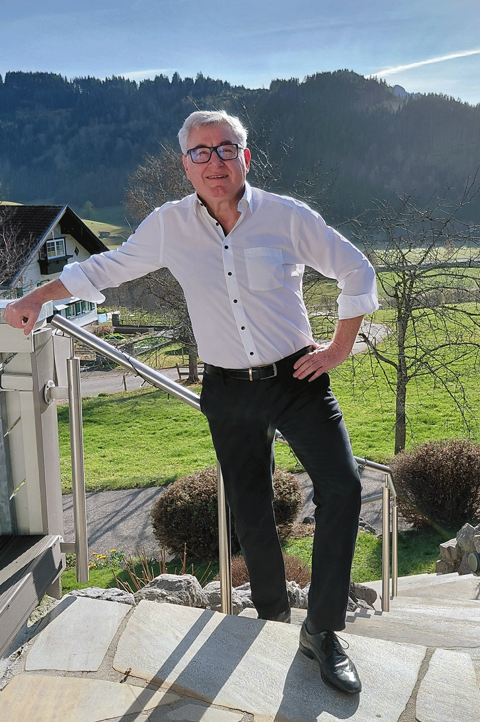 Mann mit Brille steht an einer Treppe, Hintergrund aus grünen Wiesen und Bergen.